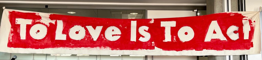 Large fabric banner in Camden Council office featuring the words 'To Love Is To Act' with a bright red painted background.