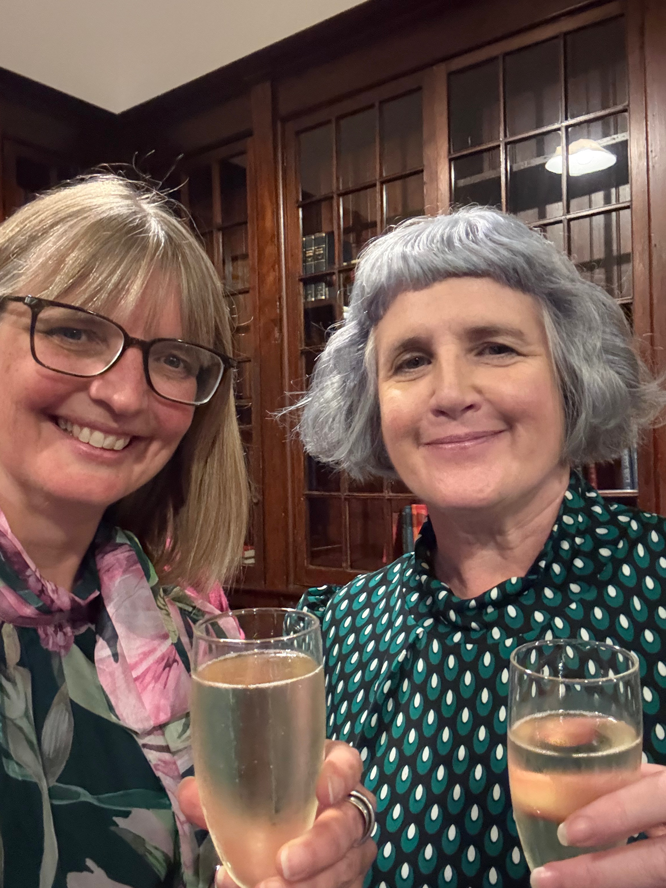 Bryony and Beverley Ward standing in front of bookshelves with glass doors, holding glasses of prosecco