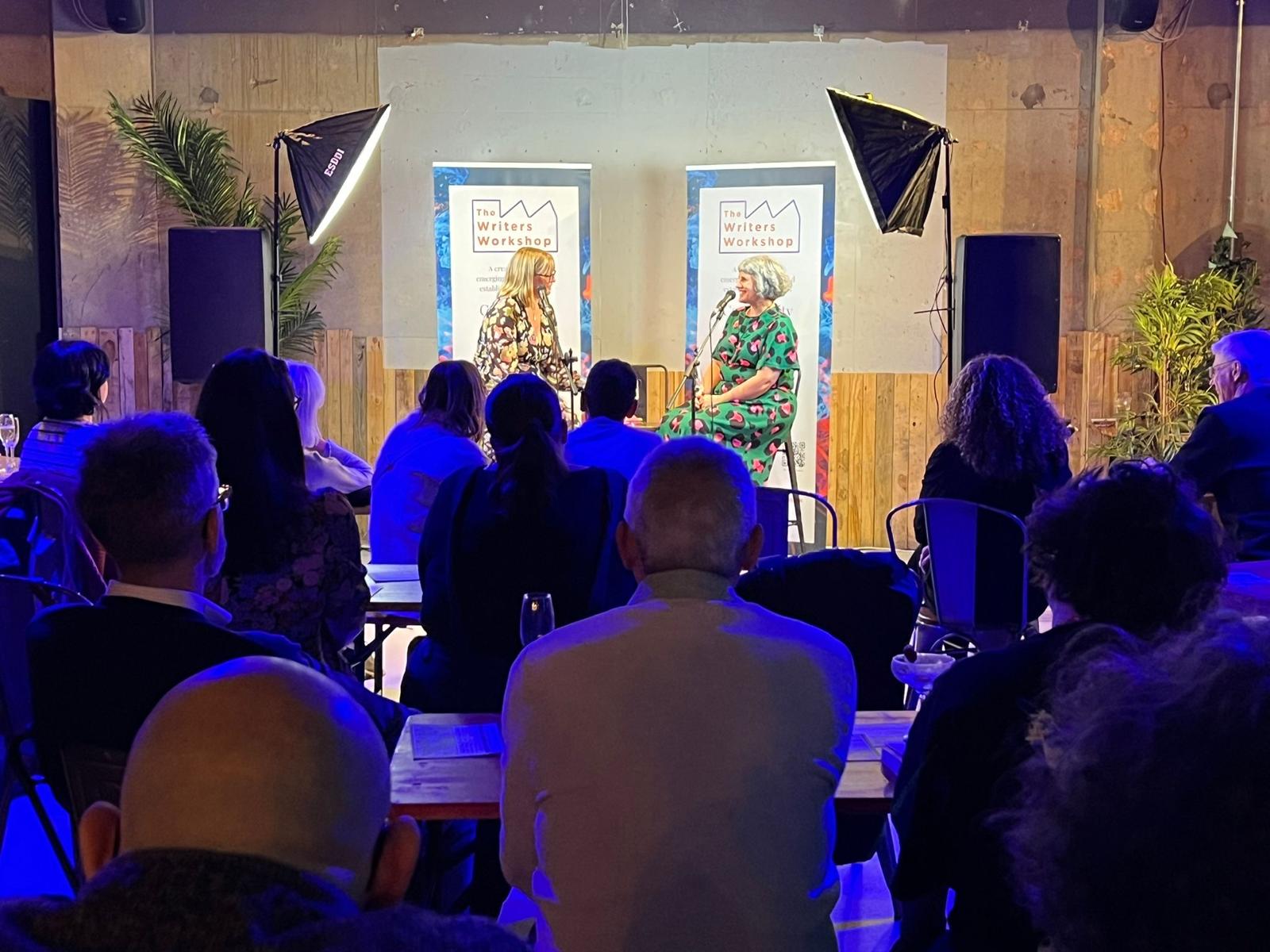 Bryony and Beverley talking at the front of a room full of people. They have big lights shining on them and they are sitting on stools in front of banners saying 'The Writers Workshop'